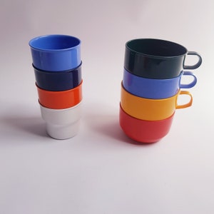 May include: A stack of six colourful plastic cups, with the top cup being blue, the second cup being dark blue, the third cup being orange, the fourth cup being white, the fifth cup being yellow, and the bottom cup being red. The cups are stacked in a pyramid shape.