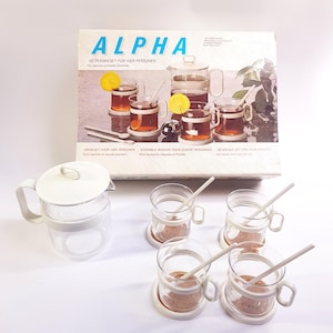 May include: A clear glass beverage set with a white lidded pitcher and four mugs with white bands and matching coasters. The set is packaged in a box with the text "ALPHA GETRANKESET FOR VIER PERSONEN" printed on it.