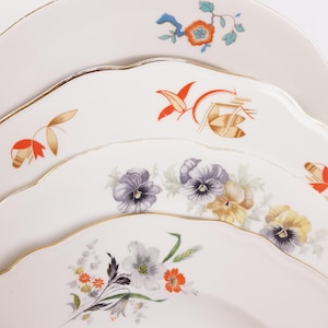 Op de afbeelding: Een stapel van vier witte keramische borden met geschulpte randen en een gouden bies. Elk bord heeft een uniek bloemmotief, waaronder oranje bloesems, abstracte oranje en gouden botanische motieven, paarse en gele viooltjes en wilde bloemen.