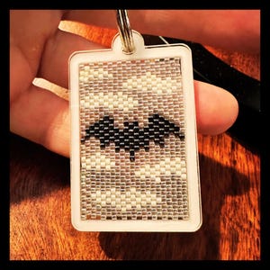 May include: A rectangular keychain featuring a beaded insert with a black bat silhouette centered against a background of white and translucent glass beads. The beaded panel is encased in a white plastic frame with a metal ring at the top.