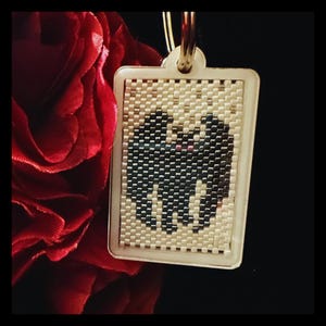 May include: A rectangular keychain featuring a beaded insert with a black bat silhouette against a cream-colored background. The bat has two small pink dots for eyes. The keychain is encased in a light-colored frame with a metal ring at the top.