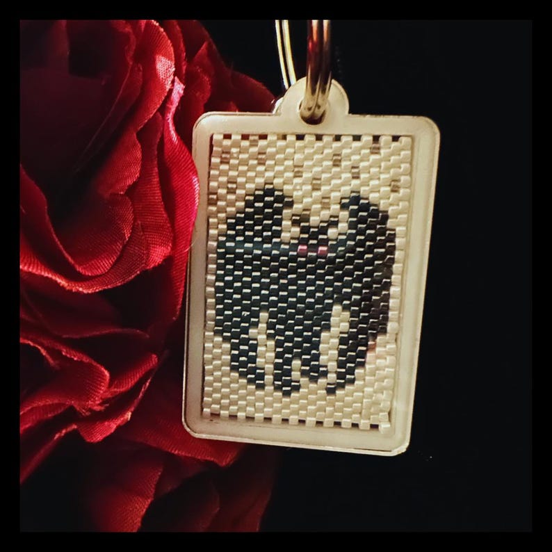 May include: A rectangular keychain featuring a beaded insert with a black bat silhouette against a cream-colored background. The bat has two small pink dots for eyes. The keychain is encased in a light-colored frame with a metal ring at the top.