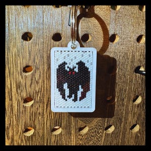 May include: A rectangular keychain featuring a beaded design of a black bat with small red eyes against a white background. The beaded piece is set within a white frame and attached to a silver metal split ring for keys.