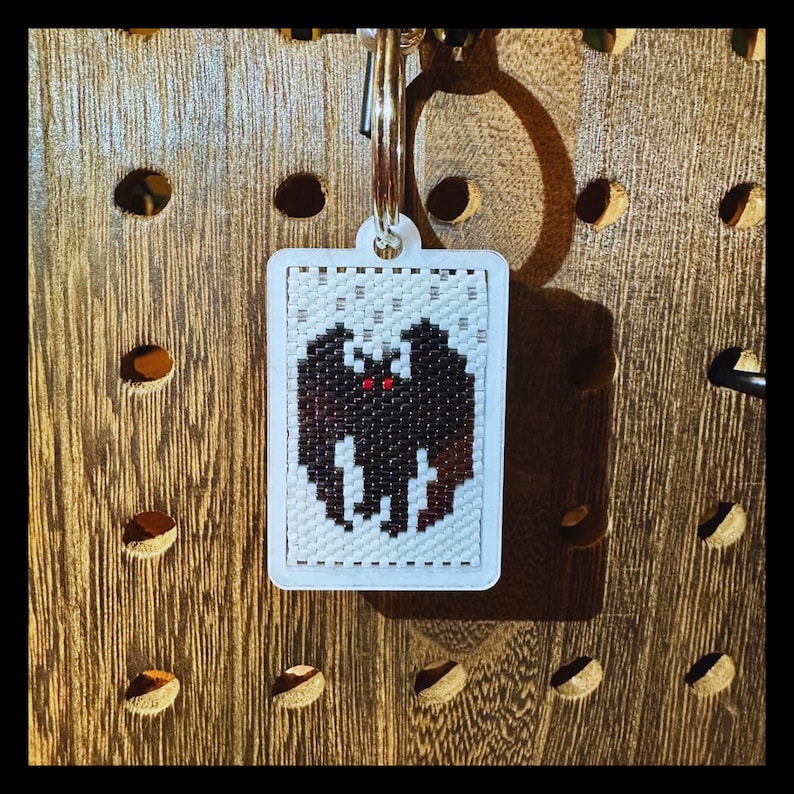 May include: A rectangular keychain featuring a beaded design of a black bat with small red eyes against a white background. The beaded piece is set within a white frame and attached to a silver metal split ring for keys.