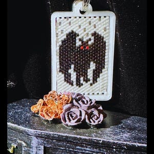 May include: A rectangular beaded pendant featuring a black bat with red eyes on a cream background, suspended from a metal ring. Below the pendant sits a small black coffin-shaped box adorned with a cluster of orange and purple miniature roses.