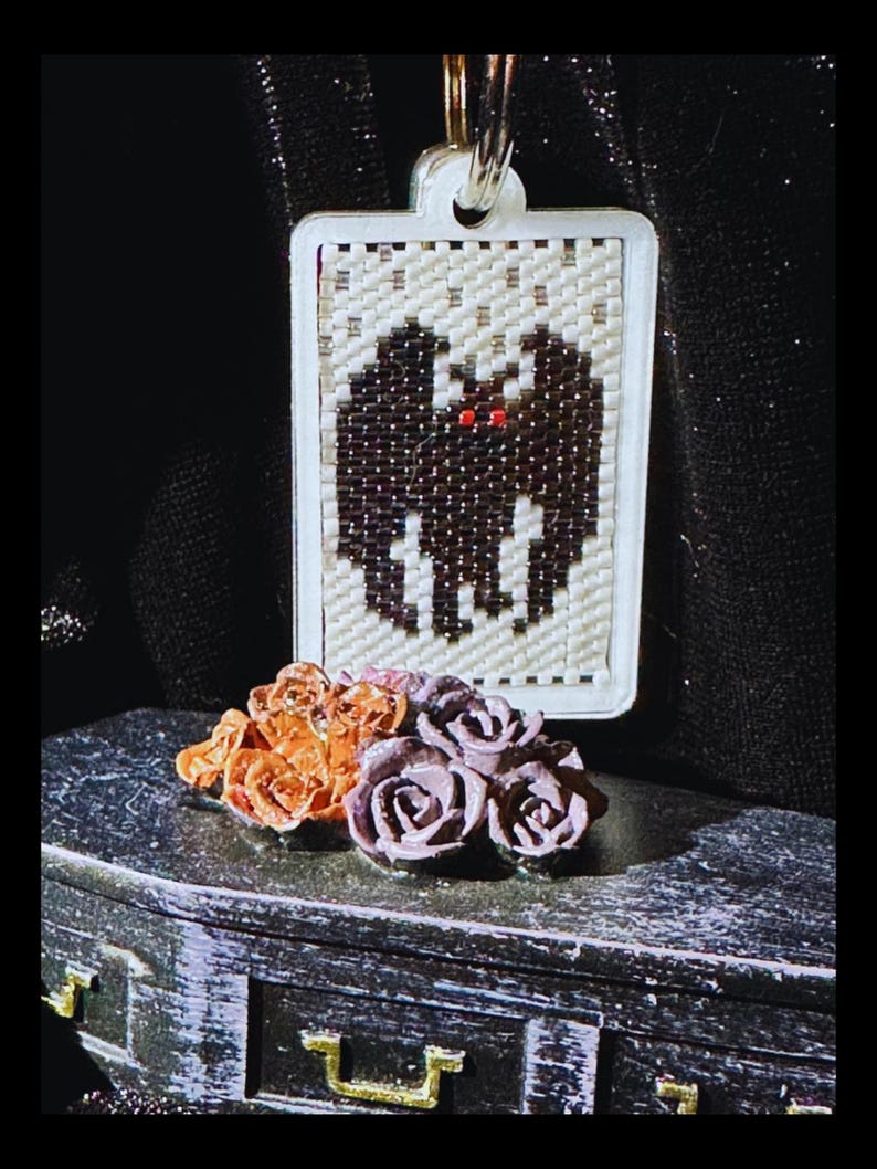 May include: A rectangular beaded pendant featuring a black bat with red eyes on a cream background, suspended from a metal ring. Below the pendant sits a small black coffin-shaped box adorned with a cluster of orange and purple miniature roses.