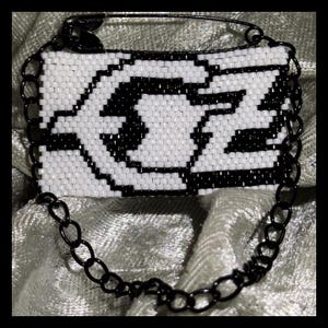 May include: A rectangular beaded patch featuring a black and white geometric pattern. The patch is attached to a black metal safety pin at the top and is connected to a black metal chain that loops around the bottom of the beaded rectangular piece.