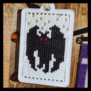 May include: A rectangular pendant featuring a beaded design of a black bat with two small red eyes centered on a white background. The beaded artwork is encased in a white plastic frame with a small loop at the top for hanging or attachment.