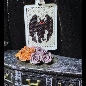 May include: A rectangular pendant featuring a beaded bat design with black wings and red eyes on a cream background. Below the pendant sits a miniature black coffin-shaped box adorned with a cluster of small orange and purple sculpted roses.