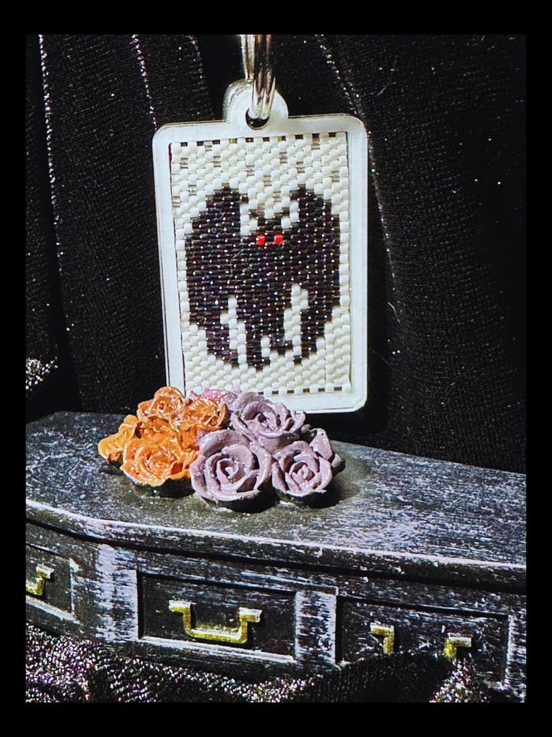 May include: A rectangular pendant featuring a beaded bat design with black wings and red eyes on a cream background. Below the pendant sits a miniature black coffin-shaped box adorned with a cluster of small orange and purple sculpted roses.