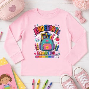 May include: A light pink long-sleeve shirt featuring a graphic print of a colorful backpack filled with school supplies, surrounded by crayons and an apple. The text reads "KINDERGARTEN LOADING..." above a progress bar with colorful segments.