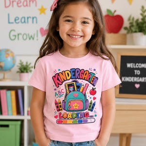 May include: A light pink short-sleeved t-shirt featuring a colorful graphic print of a teal backpack, school supplies, and the text "KINDERGARTEN LOADING" with a rainbow-colored progress bar at the bottom, worn with blue denim jeans.