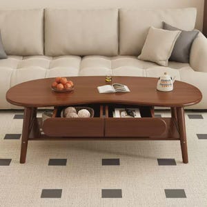 Op de afbeelding: Een niervormige houten salontafel met een gladde bruine afwerking, voorzien van twee open lades met decoratieve bollen en een boek. De tafel heeft vier taps toelopende poten en een onderste plank, met daarop een schaal met sinaasappels en een theepot.