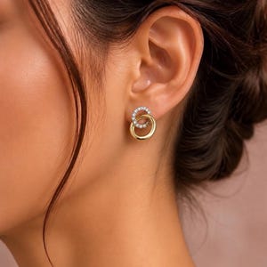 May include: A stud earring featuring two interlocking circular rings. The top ring is encrusted with small, clear, sparkling stones, while the bottom ring is a smooth, polished gold-tone metal. The design creates a layered, circular effect.