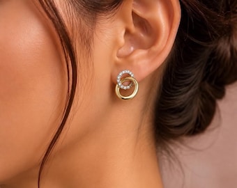 Pode incluir: Um brinco de pressão com dois anéis circulares entrelaçados. O anel superior é incrustado com pequenas pedras claras e brilhantes, enquanto o anel inferior é de metal dourado liso e polido, criando um efeito circular em camadas.