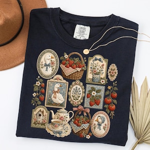 May include: A dark navy blue t-shirt featuring a graphic print of various framed illustrations including geese, baskets of strawberries, a teapot with a teacup, and floral arrangements. The shirt has a Comfort Colors brand label at the inner neckline.