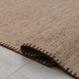 May include: A rectangular area rug featuring a textured, light brown woven surface with a speckled appearance. The edges of the rug are finished with a dark brown whipstitch detail, creating a visible border along the folded corner of the fabric.