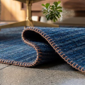 May include: A deep blue area rug with a textured, ribbed surface and a folded corner. The edges of the rug are finished with a thick, tan-coloured whipstitch detail that runs along the entire perimeter, providing a distinct contrast to the blue.