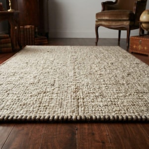 May include: A rectangular area rug featuring a thick, hand-woven texture with a chunky, looped pile construction. The rug displays a neutral color palette of off-white, cream, and light beige tones, finished with a dark brown braided edge border.