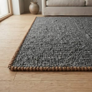 Könnte beinhalten: Ein rechteckiger Teppich mit einer dicken, dunkelgrauen Strickstruktur. Der Teppich ist mit einer markanten, geflochtenen beigen Jutekante versehen, die entlang des gesamten Umfangs verläuft und einen deutlichen Kontrast zur grauen Mitte bildet.