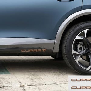 Op de afbeelding: Een mat donkerblauw zijpaneel van een auto met een onderste grijze sierstrip met de koperkleurige tekst CUPRA en een bijpassend logo. Het voertuig heeft een zwart-zilveren lichtmetalen velg met een koperen naafdop. Twee extra CUPRA-stickers.