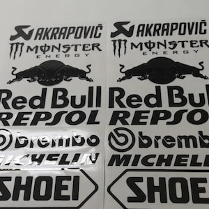 Peut inclure: Deux feuilles d'autocollants verticales identiques présentant des logos et du texte noirs sur fond blanc. Les motifs incluent les marques Akrapovic, Monster Energy, Red Bull, Repsol, Brembo, Michelin et Shoei, ainsi qu'un graphique de deux taureaux.