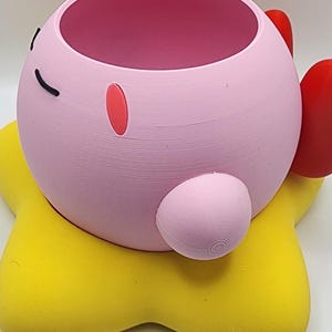 May include: A spherical pink planter featuring a smooth surface, a dark pink interior, and small black eyes. The planter is shaped like a character with a small pink oval cheek, a round arm, and is resting on a bright yellow star-shaped base.
