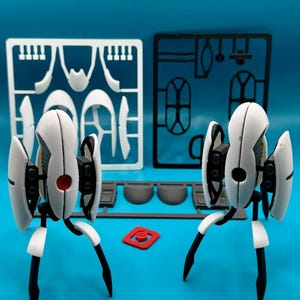 May include: Two white and black robotic turret figures with three legs and a central eye. One turret has a red eye, the other a black eye. Behind them are white and black plastic model kit frames, a grey plastic component, and a small red square piece.