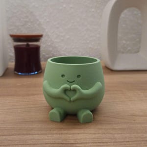 May include: A light green, rounded planter featuring a small, smiling character with arms positioned to form a heart shape in front of its body. The planter has two small feet at the base and a hollow, open top for holding small items or plants.
