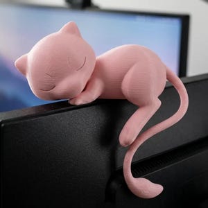 May include: A light pink, smooth-textured figurine of a small, cat-like creature with pointed ears and closed eyes, depicted in a sleeping position. The figure is designed to rest securely on the top edge of a black computer monitor screen.