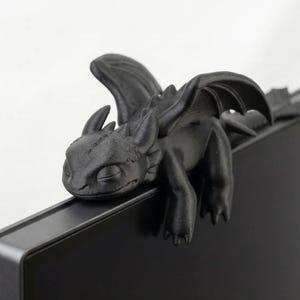 Może przedstawiać: Matowa czarna figurka smoka wydrukowana w 3D z dużymi skrzydłami, spiczastymi uszami i gładką teksturą, zaprojektowana tak, aby siedziała na górnej krawędzi płaskiego monitora. Stworzenie ma zaokrągloną głowę, zamknięte oczy i długi ogon z tyłu.