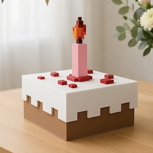 May include: A square, pixelated-style cake box with a brown base and white top featuring a jagged edge. A pink candle with a multi-colored orange and red flame sits on the center of the lid, surrounded by small red square decorative accents.