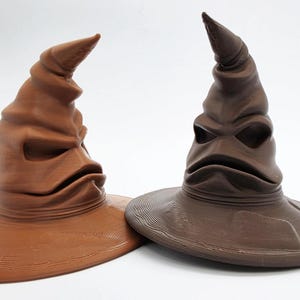 May include: Two pointed hats with wide brims and textured, folded surfaces. One hat is a light reddish-brown, while the other is a darker, deep brown. Both feature a distinct, curved top and a wide, expressive mouth-like opening near the base.