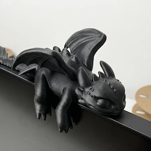 May include: A matte black 3D printed dragon figurine with a smooth texture, featuring large wings, pointed ears, and a long tail. The creature is posed in a resting position, with its front limbs and head draped over the top edge of a black monitor.