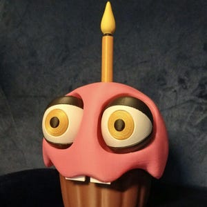 Puede incluir: Una figura de cupcake rosa con base marrón, que presenta dos ojos grandes con esclerótica blanca e iris dorados, dos dientes blancos pequeños y una vela amarilla única que sobresale del centro superior de la superficie rosa.