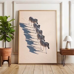 Op de afbeelding: Een ingelijste kunstdruk met een rij van vijf zebra's die achter elkaar over een lichtbeige, geribbelde zandduin lopen. Elke zebra heeft een opvallend zwart-wit streeppatroon en werpt lange, donkere schaduwen op het zand.