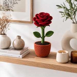 Könnte beinhalten: Eine handgefertigte Häkelrose mit einer leuchtend roten Blüte aus geschichteten Blütenblättern, einem grünen Stiel mit zwei Blättern und einer dunkelbraunen Erde, alles in einem strukturierten, runden, terrakottafarbenen Häkeltopf.