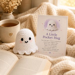 Op de afbeelding: Een wit gehaakt spookfiguurtje met zwarte ogen en roze wangen, vergezeld van een rechthoekige kaart met een spookillustratie en de tekst: A Little Pocket Hug. This little ghost is here to remind you: even on your foggiest days, you're never really alone. Tuck it in your pocket, sit it on your desk, or give it a squeeze whenever you need a little extra comfort.