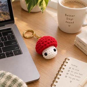 May include: A crochet mushroom keychain featuring a bright red cap with a textured surface and a cream-colored stem. The mushroom has two small black bead eyes and is attached to a gold-toned metal split ring by a short chain link connector.
