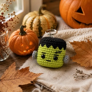 May include: A crochet keychain shaped like a green monster head with black hair, black eyes, a small mouth, and a stitched scar on the forehead. The figure has small grey bolts on the sides and a silver metal keyring attached to the top.