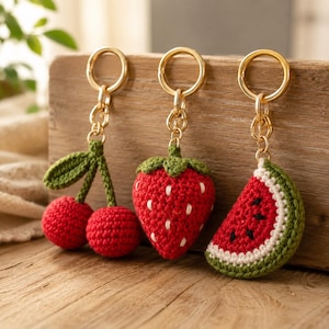 May include: Three crochet fruit keychains featuring a pair of red cherries with green leaves, a red strawberry with white seed details, and a red watermelon slice with a white rind and green edge, each attached to a gold-toned metal ring.