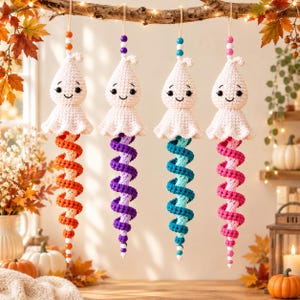 May include: Four crocheted ghost ornaments hanging in a row, each featuring a white ghost head with a smiling face and a long, spiraling tail in orange, purple, teal, or pink. Each ornament is topped with a string of colorful wooden beads.