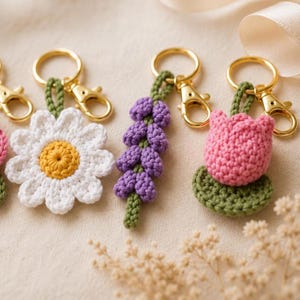 May include: Three crochet keychains featuring floral designs: a white daisy with a yellow center, a purple lavender sprig, and a pink tulip on a green leaf. Each keychain is attached to a gold-toned metal lobster clasp and a circular ring.