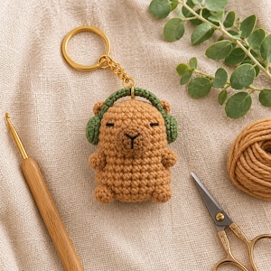 Peut inclure: Un porte-clés capybara en crochet beige avec des cache-oreilles vert foncé, un anneau en métal doré et une chaînette. Le capybara présente des yeux et un nez brodés en noir, avec une surface en laine texturée. L'objet est présenté avec un crochet en bois, des ciseaux et une pelote de laine beige.