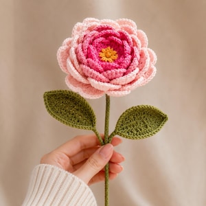 May include: A handcrafted crochet flower featuring layered petals in shades of light pink and vibrant magenta, with a small yellow center. The flower is attached to a thin green stem with two textured, pointed green leaves extending from the sides.