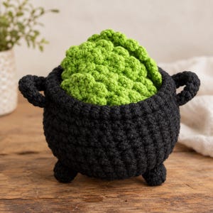 May include: A black crocheted cauldron with two side handles and three small feet, containing several bright green crocheted circular coasters with a textured, bumpy surface that mimics the appearance of bubbling liquid or foam.