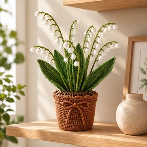 May include: A crocheted lily of the valley plant featuring white bell-shaped flowers and long green leaves. The plant is potted in a textured brown crocheted container that is decorated with a small, tied bow made from the same brown yarn.