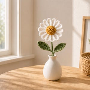 May include: A handcrafted crochet daisy flower with white petals and a textured yellow center, attached to a green stem with two green leaves. The flower is displayed in a small, smooth, matte white ceramic vase with a rounded base and narrow neck.