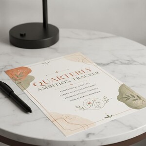 May include: A white paper sheet titled Quarterly Ambition Tracker with professional goal log, career milestone organizer, business growth template, and goal tracking printable text. It features abstract terracotta and sage green shapes.