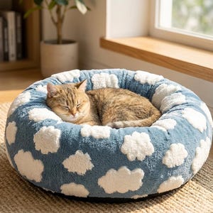 May include: A round, plush pet bed featuring a soft, blue fabric base decorated with a repeating pattern of white, fluffy clouds. A brown tabby cat is curled up and sleeping inside the center of the circular, cushioned pet bed.